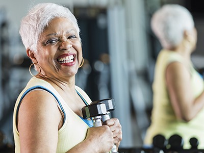 Woman in her 60s working out at the gym, staying in shape. She is smiling at the camera, holding a dumbbell in each hand, in front of a rack of weights.